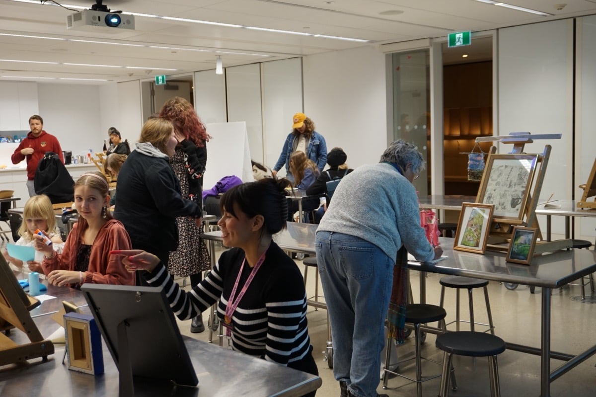 Youth Art Night participants gather in the studio to present artwork created throughout the year. Framed drawings, paintings, and other creative projects are displayed on tables and easels as attendees view and discuss the students' work.