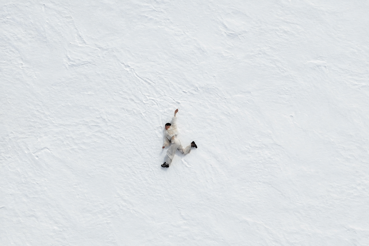 A person dressed in light-coloured winter clothing lies stretched out on a vast snow-covered landscape. Viewed from above, the figure appears small against the expansive white surroundings, emphasizing the scale and solitude of the scene.