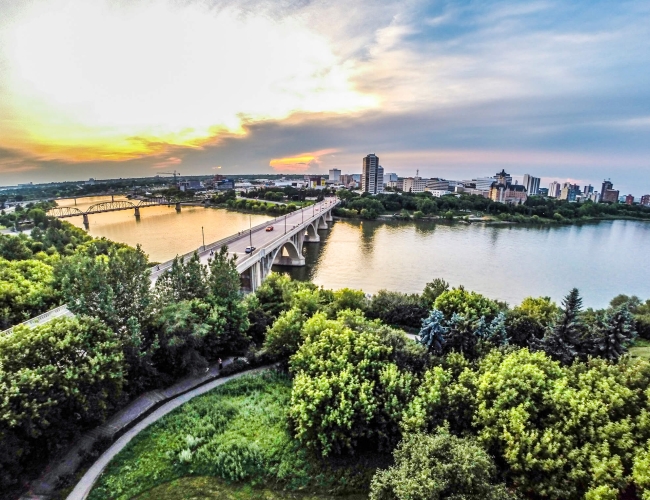 Aerial shot of Saskatoon with a green park, bridge, and water in the foreground