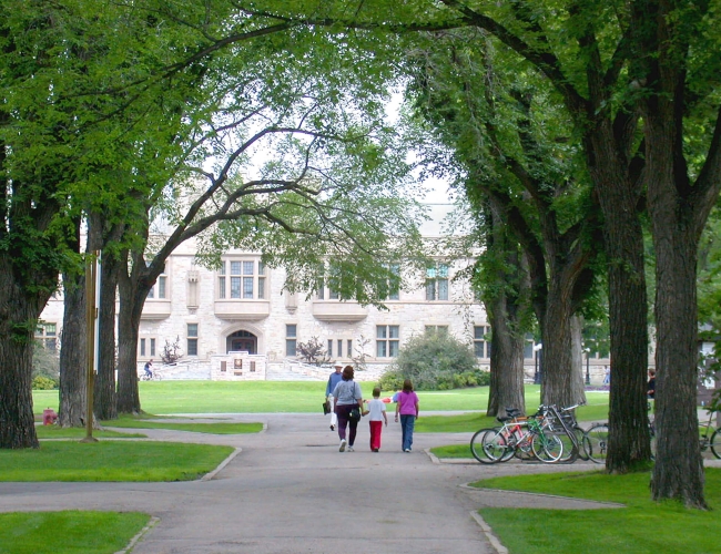 A family walking on a path underneath trees with the University of Saskatchewan main buliding in the background