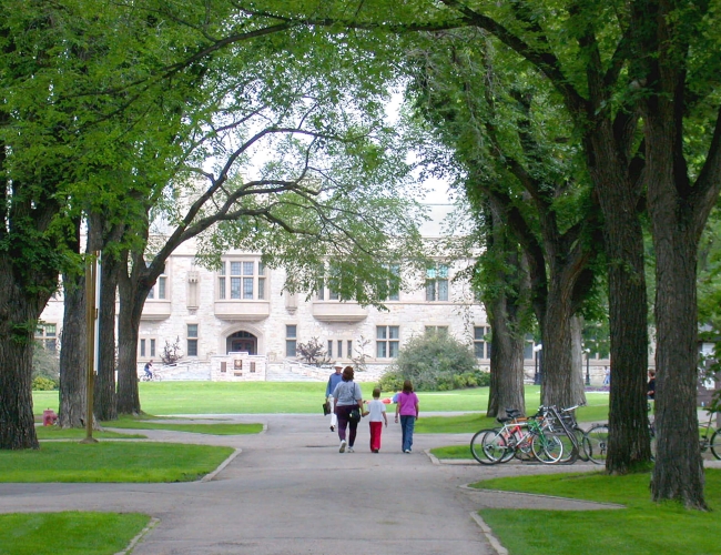 A family walking on a path underneath trees with the University of Saskatchewan main buliding in the background
