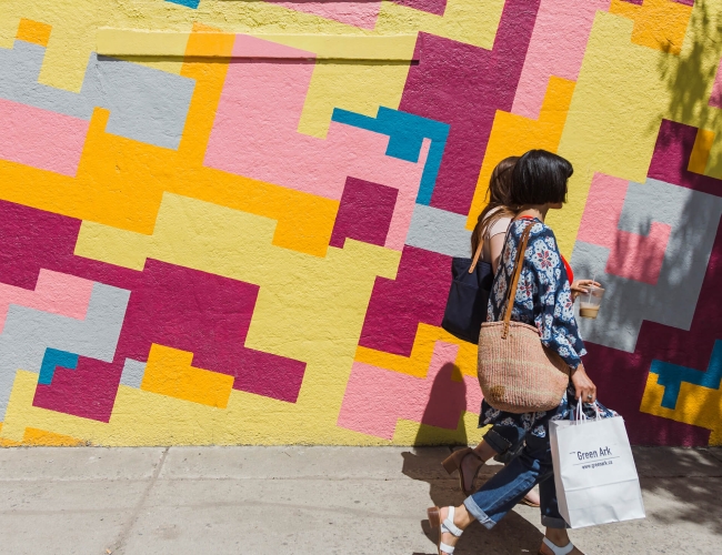two women holding shopping bags walking in front of a large wall painted with a bright mural. 