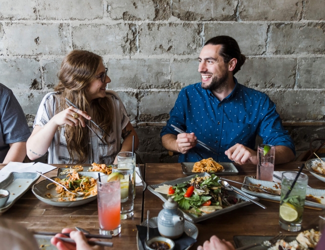 Couple sitting in front of a grey brick wall, at a wooden dining table eating