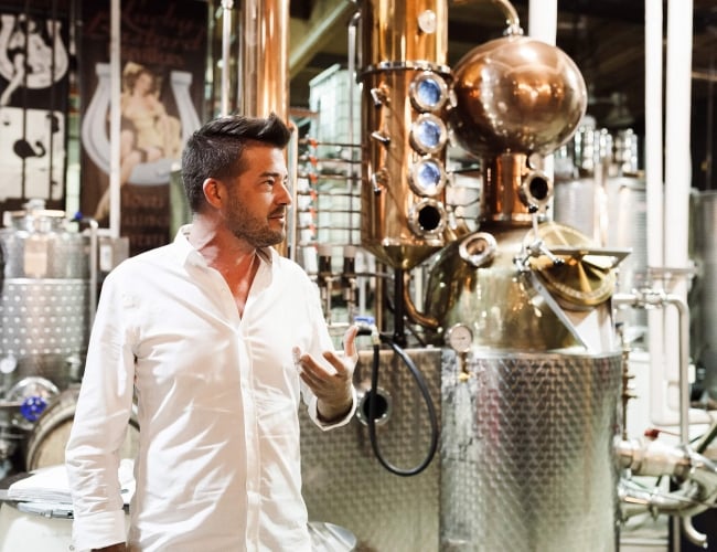 a man in a white shirt standing in front of a beer distiller, giving a tour to guests. 