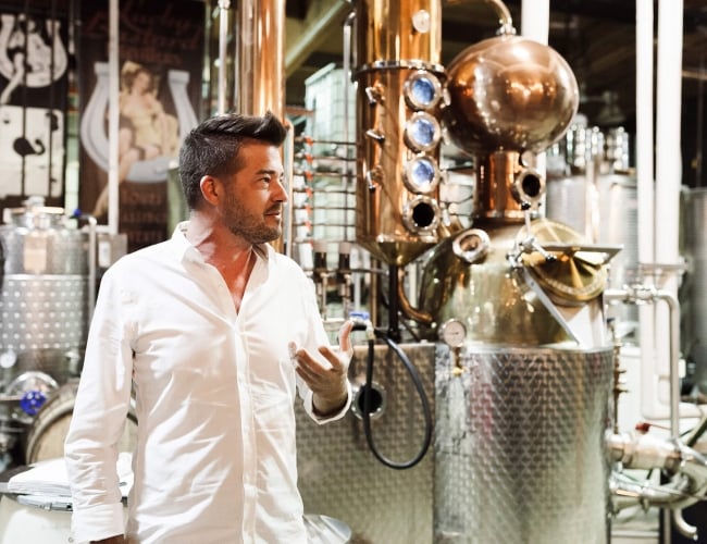 a man in a white shirt standing in front of a beer distiller, giving a tour to guests.