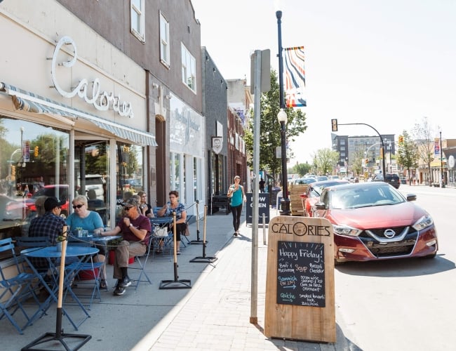 A red car parked on the side of a street lined with stores that have patio tables out front