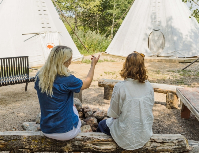 two women sitting on a bench looking at a teepee