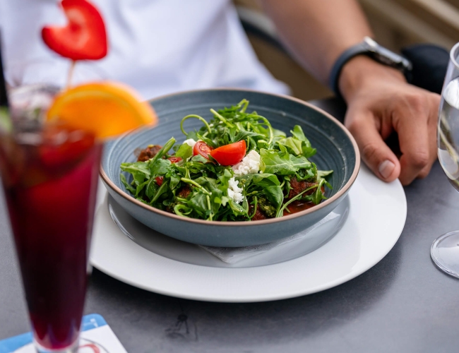 an individual sitting at an outdoor table eating a salad and drinking a cocktail