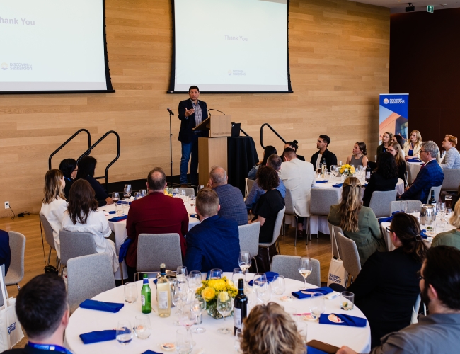 A male keynote speaker on a stage addressing a large room filled with individuals. Behind her is a Power Point Presentation.