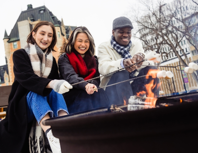 Three individuals sitting around an outdoor fire pit roasting marshmallows in the winter