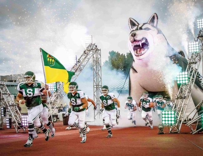 A football team runs onto the field with confetti in the air and their husky mascot behind them 