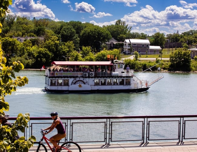 A woman ridding a bicycle in a park with a river and riverboat in the background