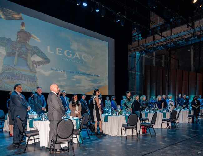 A group of individuals at a gathering standing at round tables. On the screen behind them is a slide that says "Legacy Project"