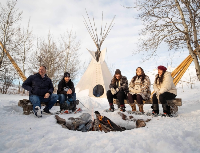 Four individuals sitting outside at wintertime in front of a fire. There is snow on the ground and a teepee in the background