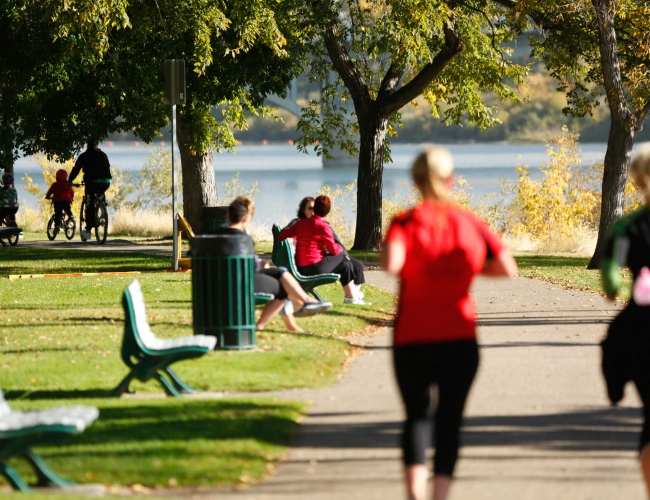 Two women running in a park. In the background some additional individuals are sitting on a park bench.