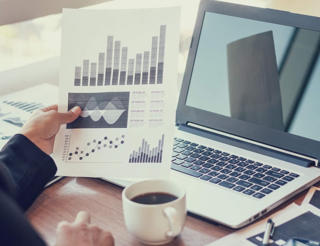 Laptop open with a spreadsheet held in front of it and cup of coffee