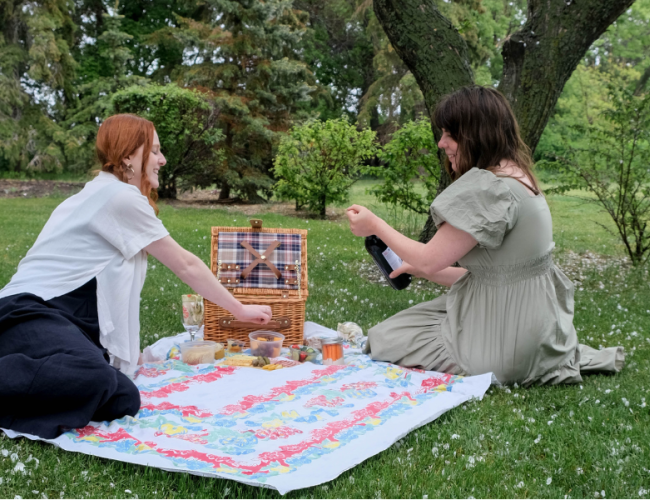 Two people sitting on a blanket, while one opens a bottle of wine.