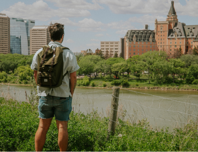 Man standing on a trail beside the river, looking at the Bessborough Hotel. 