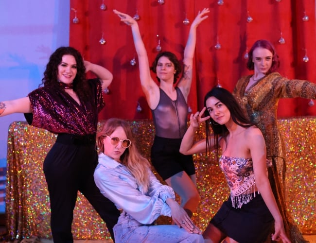 5 dancers wear 70's inspired outfits and strike disco dance poses in front of a gold glittery backdrop.