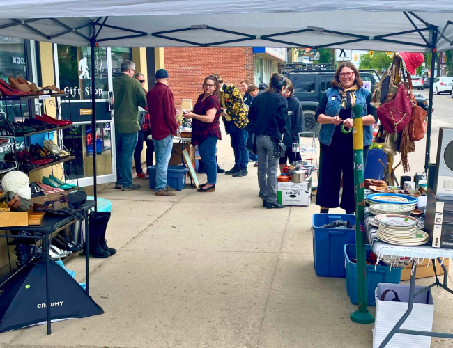 picture of people smiling and talking on the sidewalk with a market tent, shelves, and tables with goods to shop