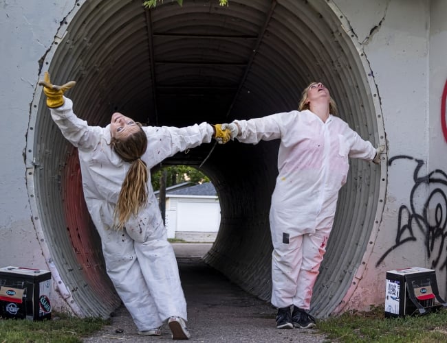 2 dancers in white paint suits hold hands and bend backwards in a metal underpass tunnel