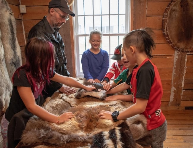 Kids playing with furs that would've been popular during the fur trade era, smiling and laughing while park interpreter smiles with them.