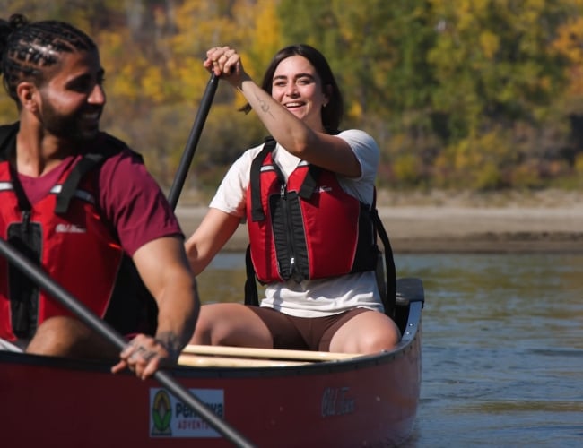 Man and women paddle in a canoe on still river smiling with fall leaves in the background