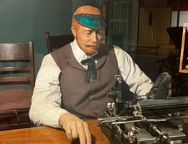 The image shows a mannequin portraying a telegrapher. He is sitting at a desk in front of a typewriter wearing a green visor and brown vest.