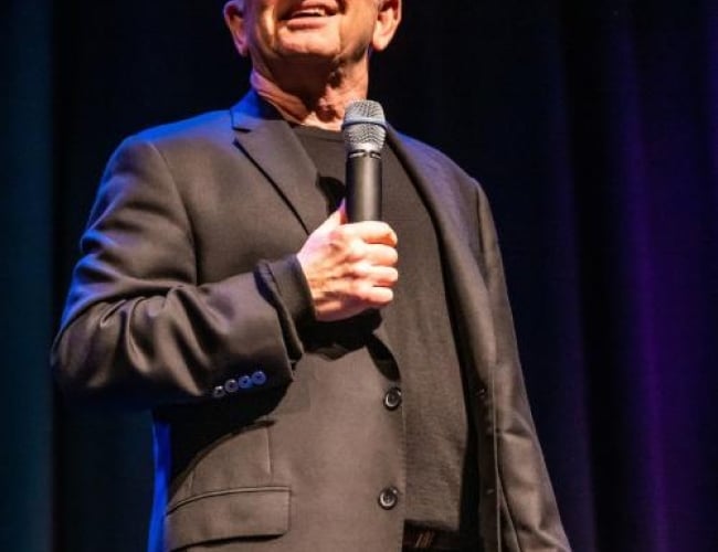 Image of comedian Ron James looking out to the audience while holding a microphone to his face. 