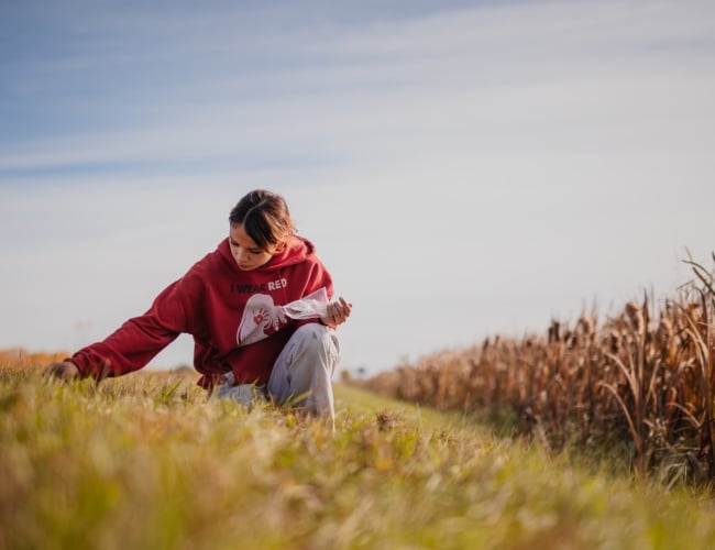 A young child wearing a red hoodie crouches in the grass looking curiously at the ground.