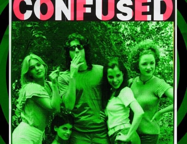 Image of some of the actors in Dazed and Confused, with one guy smoking and wearing sunglasses while theres 4 girls huddled around him, posing for a picture.