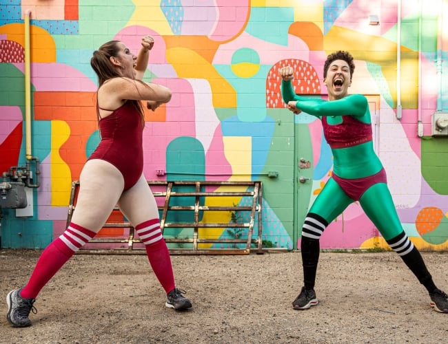 two dancers move excitedly in front of a colourful wall mural