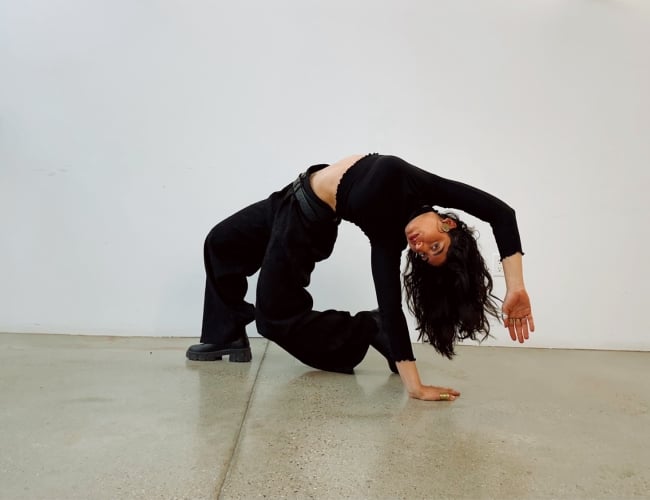 A performer (Rawda Mabrouk) dressed in black strikes a dramatic, arched pose on a polished concrete floor against a plain white wall. One hand and one knee support the body while the torso curves backward, creating a sculptural, dance-like shape.
