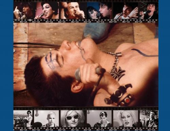 Image of a man laying down on ply wood, with his eyes closed. He is holding a mic and wearing 'punk' jewlery. There is film reel around him.