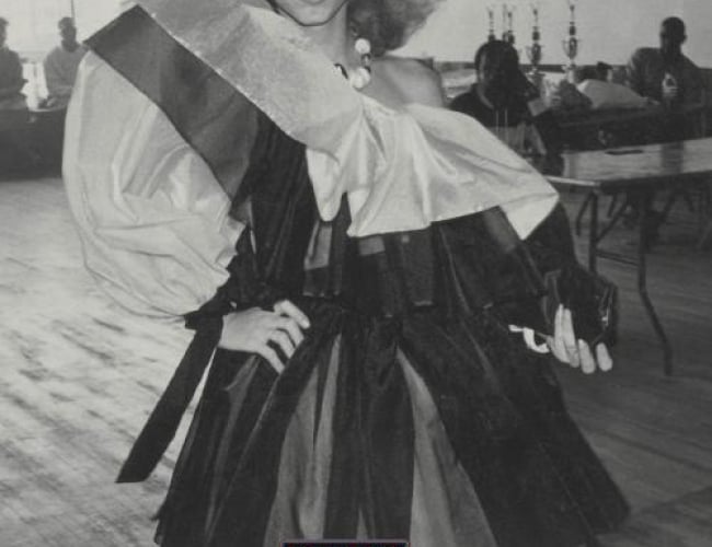 Image of someone wearing a couture dress and posing in what seems to be a ballroom. Image is in black, white and grey.