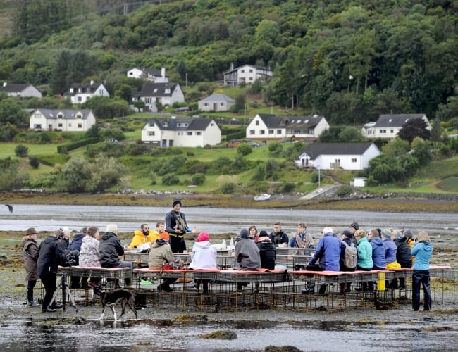 CLIMAVORE: On Tidal Zones. Public workshop by Rory MacPhee at low tide. A group of people gather around tables set on a tidal shoreline while a guide speaks at the center. Across the water, a small village and forested hillside create a scenic backdrop.