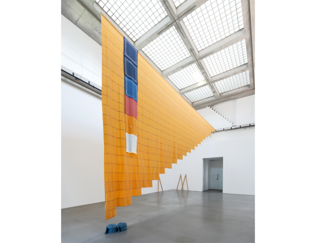 A large textile installation (Tanya Lukin Linklater, Crested, 2026) hangs from the ceiling of a bright gallery with a grid-patterned skylight. The suspended artwork features layered orange fabric with blue, red, and white elements, cascading diagonally across the open space.