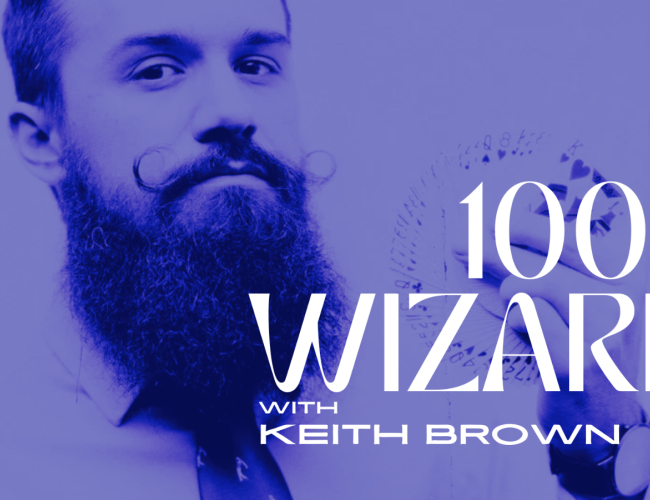 Bearded magician Keith Brown holds a fan of playing cards near his face. The entire image is in blue. There is white text that says 100% Wizard with Keith Brown.