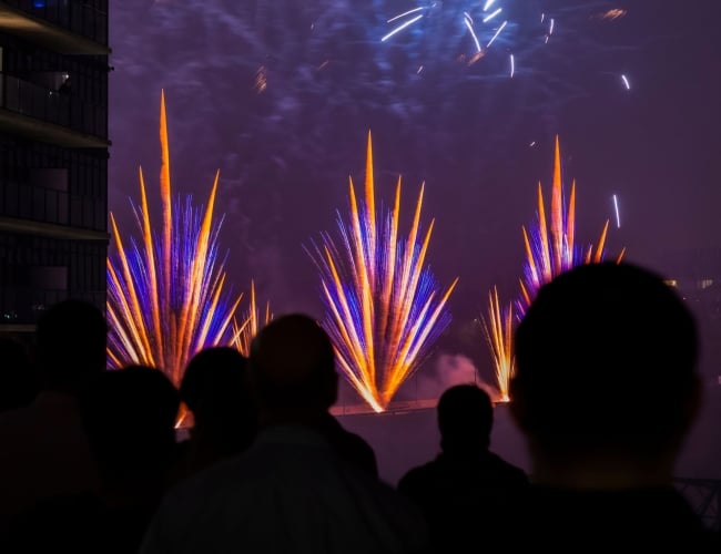 Silhouetted spectators watch a nighttime fireworks display, with bright blue and orange bursts lighting the sky. The colorful pyrotechnics rise above the water beside nearby buildings, creating a dramatic urban scene.
