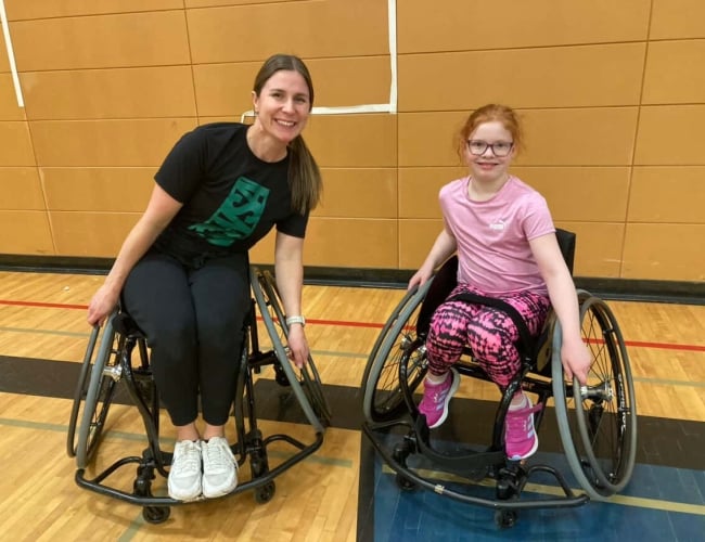 Female coach in a sport wheelchair beside a young girl in a sport chair. Both are smiling  looking at the camera and have their hands on the wheels.