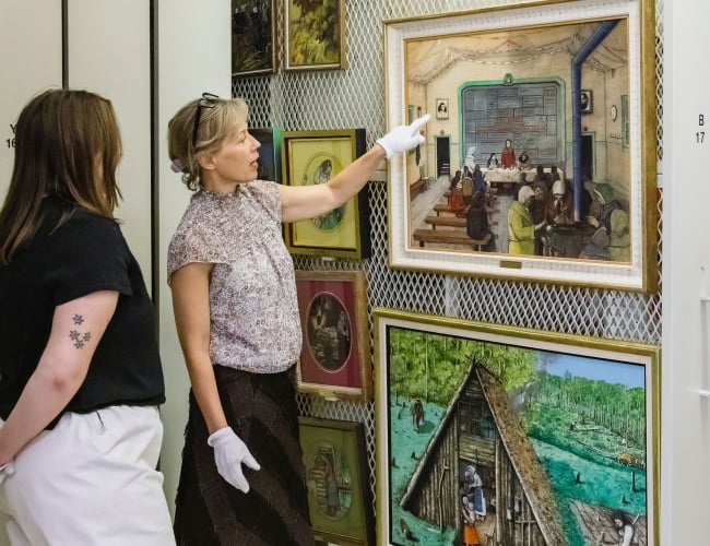 A photo of Dr. Jen Budney, Executive Director & CEO of the Ukrainina Museum of Canada, giving a tour of the collection and pointing to one of the Kurelek's paintings.