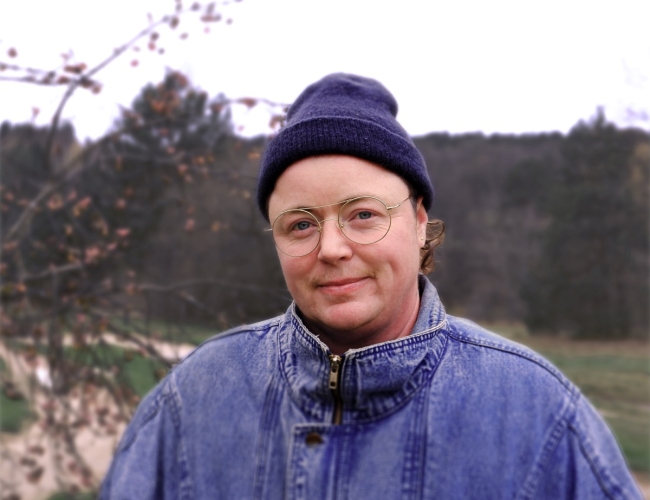 Portrait of artist Lou Sheppard standing outdoors in front of a softly blurred landscape. Lou wears round glasses, a dark knit cap, and a blue denim jacket, with bare tree branches and a wooded background visible behind him.