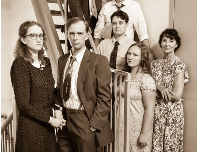 A group of actors in 1930s dress stand stoically against a steep metal staircase.