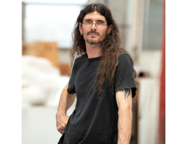 Portrait of artist, d harding, with long dark hair and glasses, wearing a black T-shirt and standing indoors. The person faces slightly to the side with one hand at their hip, while a softly blurred studio or workshop setting appears in the background.