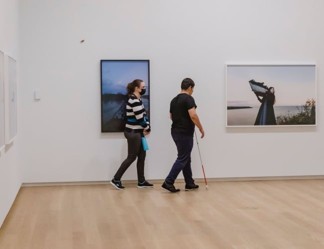 A participant uses a white cane to navigate an art gallery during a described tour while a guide walks alongside. Large framed photographs hang on the gallery walls as the group moves through the exhibition space.