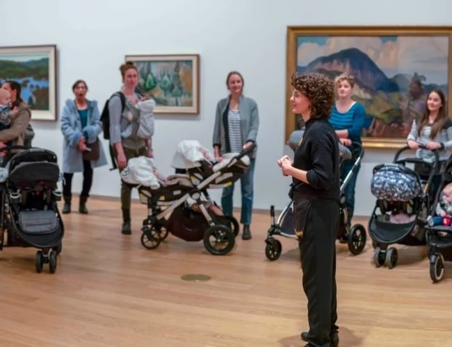 A group of caregivers and children, some in strollers, gather around a Remai Modern program guide for a tour.
