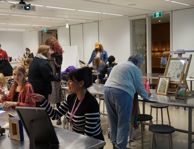 Youth Art Night participants gather in the studio to present artwork created throughout the year. Framed drawings, paintings, and other creative projects are displayed on tables and easels as attendees view and discuss the students' work.