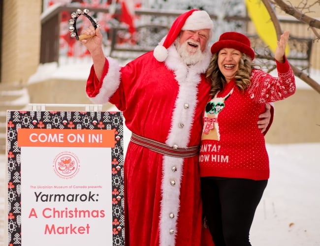 A photo of Santa and a volunteer outside of the museum next to the Yarmarok sign (sandwich board)
