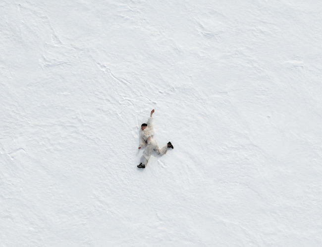 A person dressed in light-coloured winter clothing lies stretched out on a vast snow-covered landscape. Viewed from above, the figure appears small against the expansive white surroundings, emphasizing the scale and solitude of the scene.