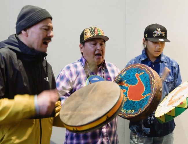 Three people stand in a circle playing hand drums and singing during a cultural performance. The drums are brightly painted with colourful designs, and motion blur captures the rhythm and movement of the drumming against a simple indoor backdrop.